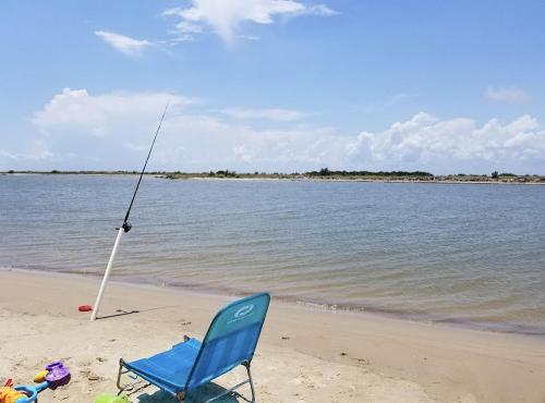 A fishing pole stands in the sand next to a blue chair by a calm waterway under a partly cloudy sky.