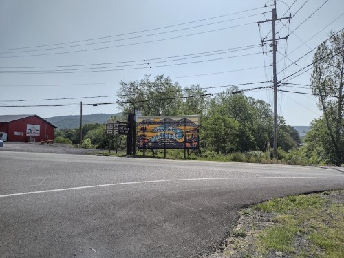 A roadside welcome sign for a town, with greenery and a red building in the background under a clear sky.