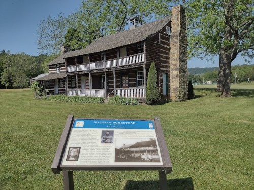 Historic wooden homestead with a stone chimney, surrounded by green grass and trees, featuring an informational sign in front.