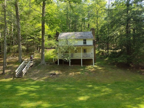 A small, two-story house surrounded by trees, with a grassy area and a staircase leading to the entrance.