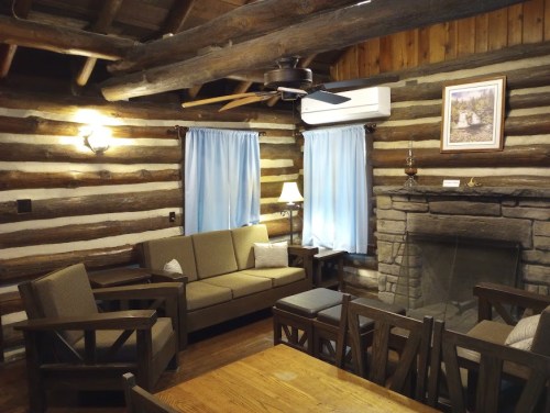 Cozy log cabin interior with wooden beams, a stone fireplace, and comfortable seating. Soft blue curtains adorn the windows.