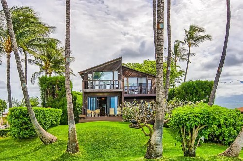 A tropical house surrounded by palm trees and lush greenery under a cloudy sky.