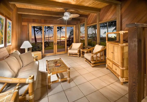 Cozy wooden living room with bamboo furniture and large windows showcasing a sunset view over palm trees.