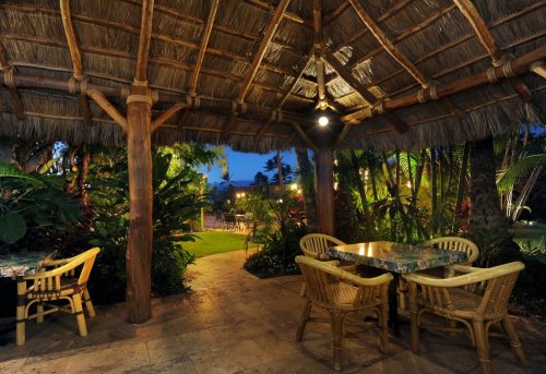 A cozy outdoor dining area with a thatched roof, surrounded by lush greenery and soft evening lighting.