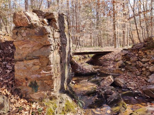 A stone structure beside a small stream, with a wooden bridge crossing over, surrounded by trees and fallen leaves.