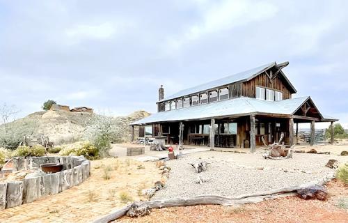 A rustic wooden house with a metal roof, surrounded by gravel and desert landscape under a cloudy sky.