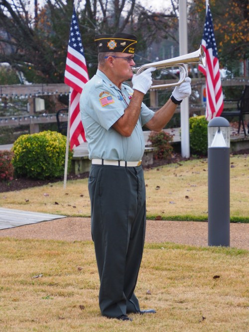A uniformed man playing a bugle outdoors, with American flags in the background and a grassy area around him.