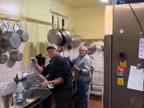 Two people wash dishes in a kitchen, surrounded by hanging pots and a stainless steel fridge.