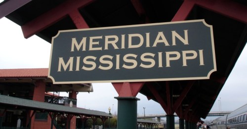 Sign at a train depot that reads Meridian, Mississippi with the town in the background.