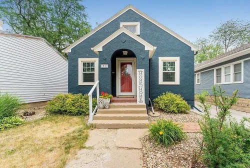 Charming blue house with a welcoming front porch and garden, surrounded by greenery and neighboring homes.