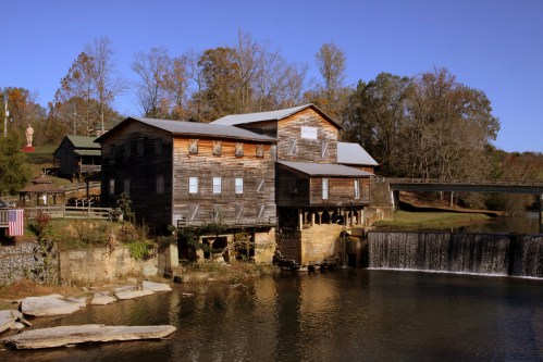 A rustic wooden mill beside a calm river, with a small waterfall and autumn trees in the background under a clear blue sky.
