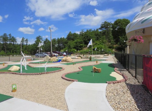 A mini-golf course with green turf, rocks, and flags under a blue sky with clouds, surrounded by trees.