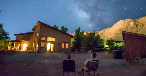 Two people sit outside a house under a starry sky, with mountains in the background and warm lights glowing from the windows.