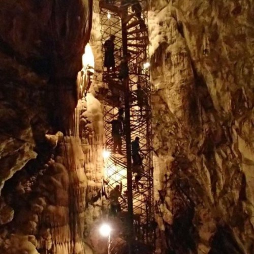 A dimly lit cave with a metal staircase and people climbing, surrounded by rock formations and illuminated by lights.