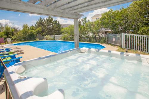 A hot tub in the foreground with a blue swimming pool and greenery in the background under a clear sky.