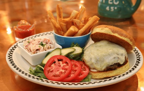 A cheeseburger with lettuce, tomato, and pickles, served with fries and coleslaw on a plate.