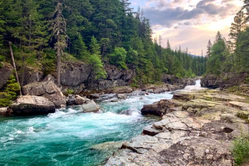 A serene river flows through rocky terrain, surrounded by lush green trees and a colorful sky at sunset.