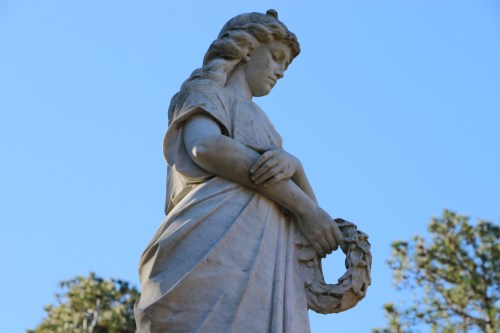 A marble statue of a woman holding a wreath, gazing downward against a clear blue sky.
