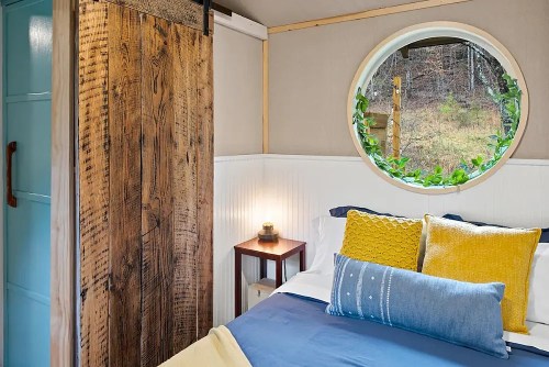 Cozy bedroom with a round window, wooden accents, and colorful pillows, surrounded by nature.