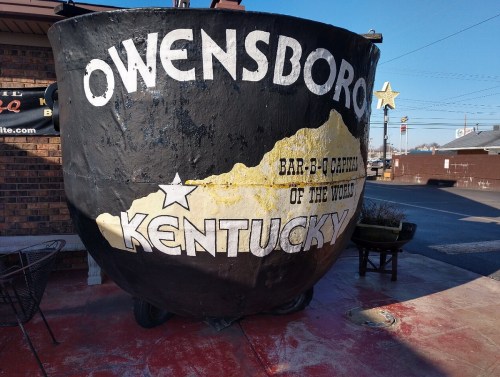 A large black pot with "Owensboro, Kentucky" and "Bar-B-Q Capital of the World" painted on it, outside a restaurant.