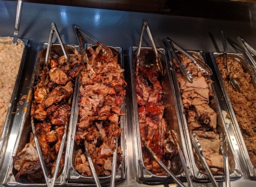 A variety of meats displayed in metal trays, with tongs for serving, showcasing a selection of barbecued dishes.