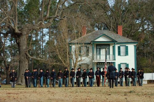 A group of soldiers in historical uniforms stands in formation in front of a large, white house surrounded by trees.
