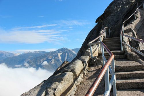 A winding staircase leads up a rocky mountain, with clouds and distant peaks visible under a clear blue sky.