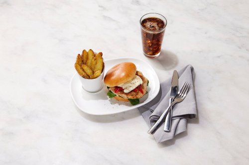 A plate with a burger topped with an egg, served with potato wedges and a glass of cola on a marble surface.