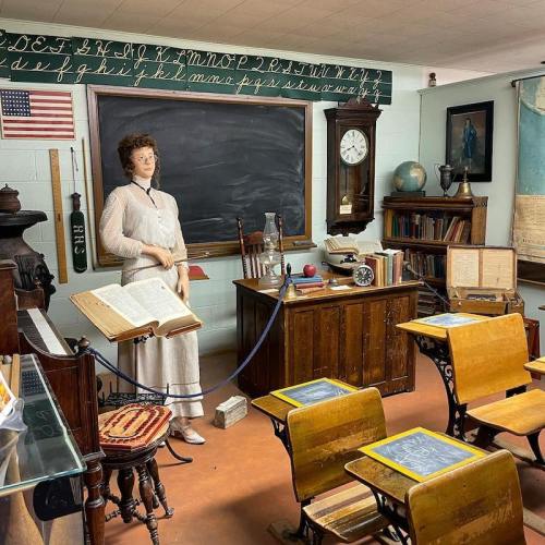 A vintage classroom scene featuring a mannequin teacher, wooden desks, a chalkboard, and historical educational materials.