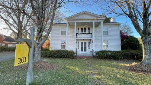 A white two-story house with a porch, surrounded by trees and greenery, featuring a yellow sign with the number 201.