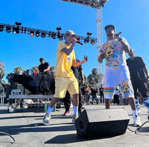 Two performers on stage, rapping into microphones, with a lively crowd and bright blue sky in the background.