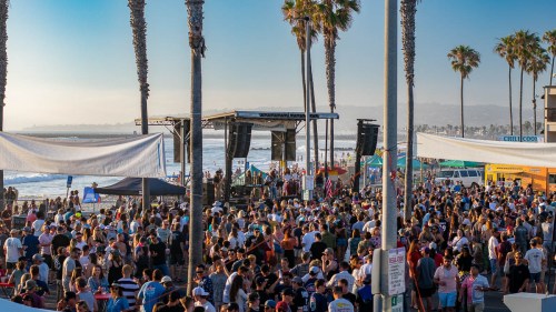 A large crowd gathers at a beach event with palm trees, a stage, and the ocean in the background.
