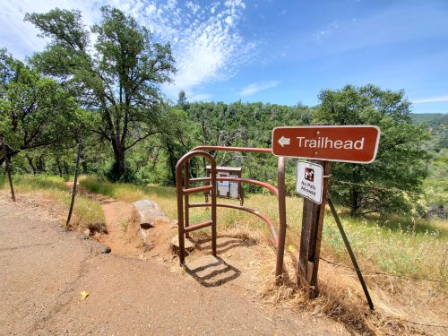 A trailhead sign with a gate, surrounded by green trees and grass under a blue sky.