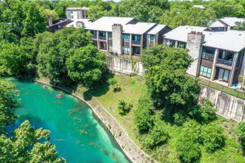 Aerial view of riverside apartments surrounded by lush greenery and a clear blue river. Kayakers visible in the water.