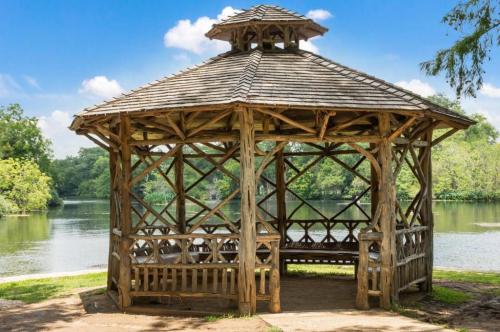 A wooden gazebo by a serene lake, surrounded by lush greenery and a clear blue sky.
