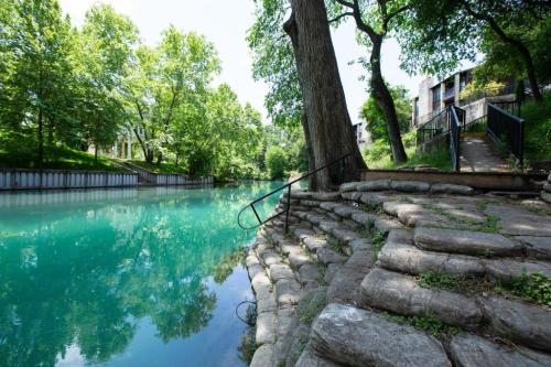 A serene riverbank with stone steps, lush green trees, and calm turquoise water reflecting the surroundings.