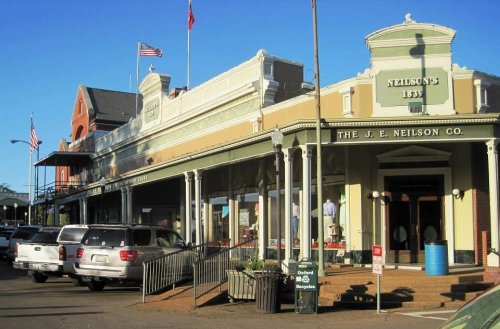 Historic storefronts with flags, featuring the J.E. Neilson Co., and parked cars in a vibrant town setting.