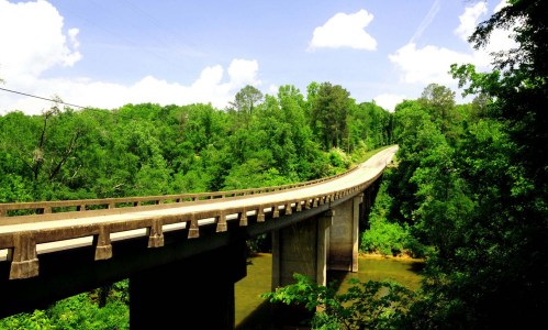 A curved concrete bridge spans a lush green landscape under a bright blue sky with fluffy white clouds.