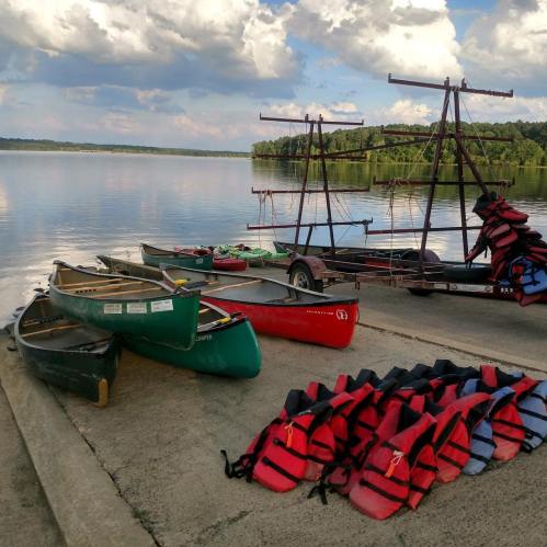 Canoes and life jackets are lined up by a calm lake under a cloudy sky, with a trailer in the background.