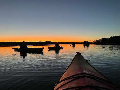 Silhouettes of kayakers on calm water at sunset, with vibrant orange and blue hues in the sky.