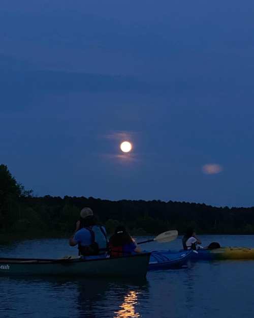 Two kayakers on a calm lake at dusk, with a bright full moon reflecting on the water.