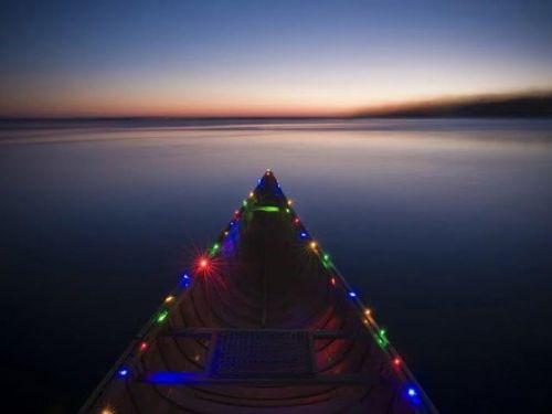 A canoe adorned with colorful lights floats on calm water at dusk, with a serene horizon in the background.