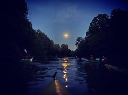 A serene night scene of kayakers on a calm river, illuminated by a full moon and surrounded by trees.