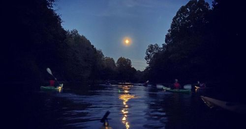 Kayakers paddle on a calm river under a full moon, surrounded by trees and a twilight sky.