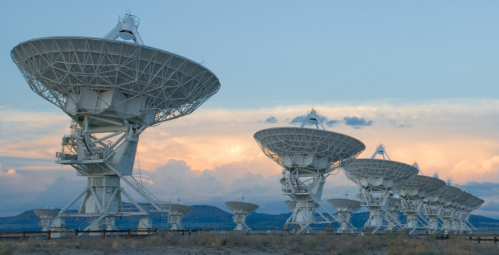 A row of large satellite dishes against a sunset sky, set in a desert landscape.