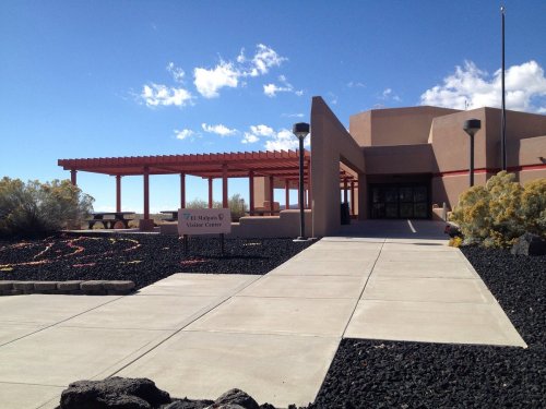 A modern visitor center with a covered patio, surrounded by black gravel and desert vegetation under a blue sky.