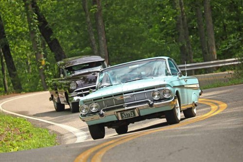 Two classic cars navigate a winding road surrounded by lush green trees.