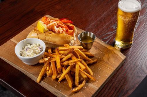 A wooden platter with a lobster roll, fries, coleslaw, and a glass of beer.