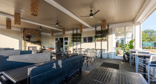 A spacious outdoor dining area with black tables, chairs, and ceiling fans, featuring greenery and a cozy atmosphere.