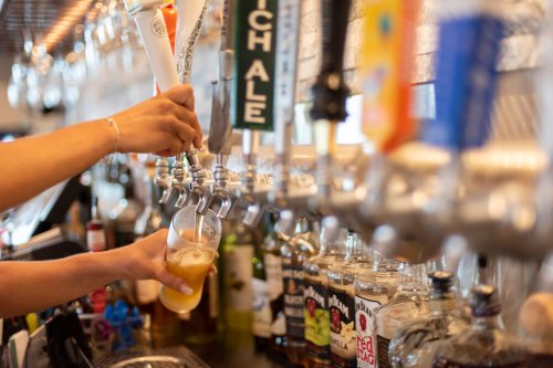 A hand pours beer from a tap into a glass, with various liquor bottles visible in the background.